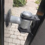 Nærbillede af et Locinox øjebolt-sæt med gevind Varmgalvaniseret hængsel på en sort port, fastgjort til stolpen. En brostensbelagt gårdhave og grønne områder ses i baggrunden.