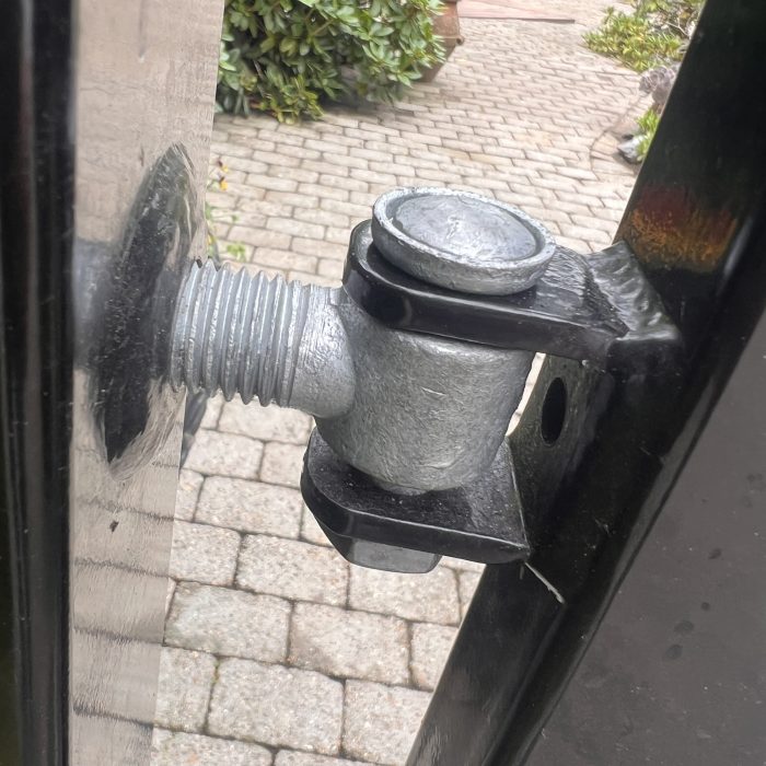 Nærbillede af et Locinox øjebolt-sæt med gevind Varmgalvaniseret hængsel på en sort port, fastgjort til stolpen. En brostensbelagt gårdhave og grønne områder ses i baggrunden.