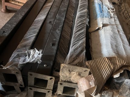 Flere "Outlet: Sorte aluminiumsstolper med fod - 70x70 mm" er stablet indendørs, nogle pakket ind i plastik og pap. Disse sorte aluminiumsstolper har flanger med firkantede åbninger i enderne.