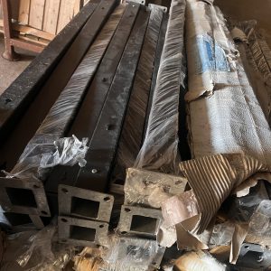 Flere "Outlet: Sorte aluminiumsstolper med fod - 70x70 mm" er stablet indendørs, nogle pakket ind i plastik og pap. Disse sorte aluminiumsstolper har flanger med firkantede åbninger i enderne.