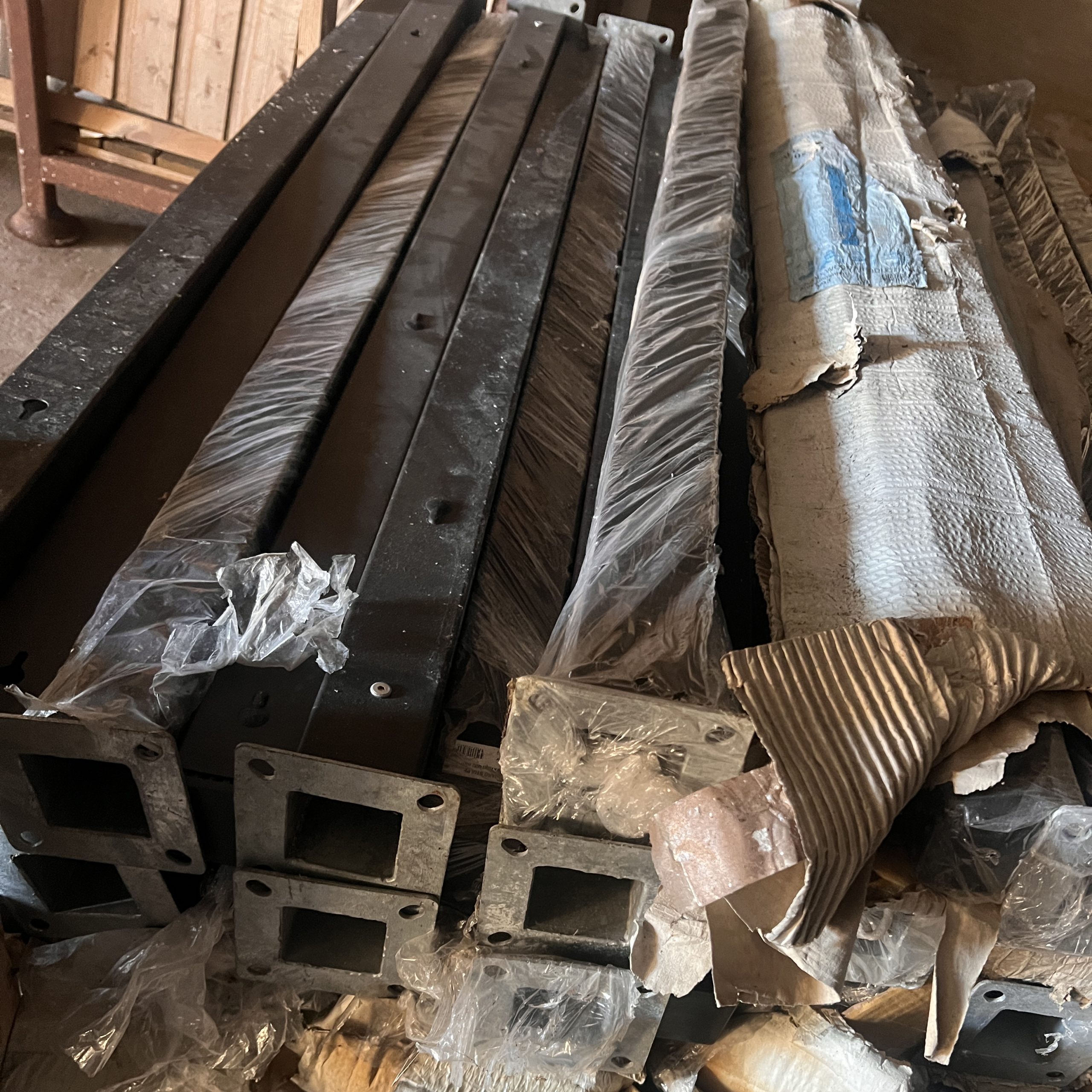 Flere "Outlet: Sorte aluminiumsstolper med fod - 70x70 mm" er stablet indendørs, nogle pakket ind i plastik og pap. Disse sorte aluminiumsstolper har flanger med firkantede åbninger i enderne.