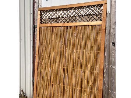 En Bambus hegn 110x170 cm træramme med en elegant gittertop læner sig op ad en trævæg under "Hegnsfabrikken"-skiltet i et lyst, solbeskinnet udendørs miljø.