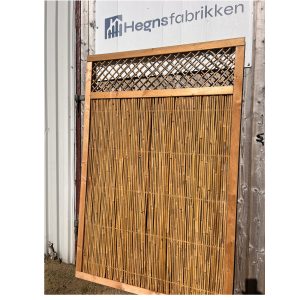 En Bambus hegn 110x170 cm træramme med en elegant gittertop læner sig op ad en trævæg under "Hegnsfabrikken"-skiltet i et lyst, solbeskinnet udendørs miljø.