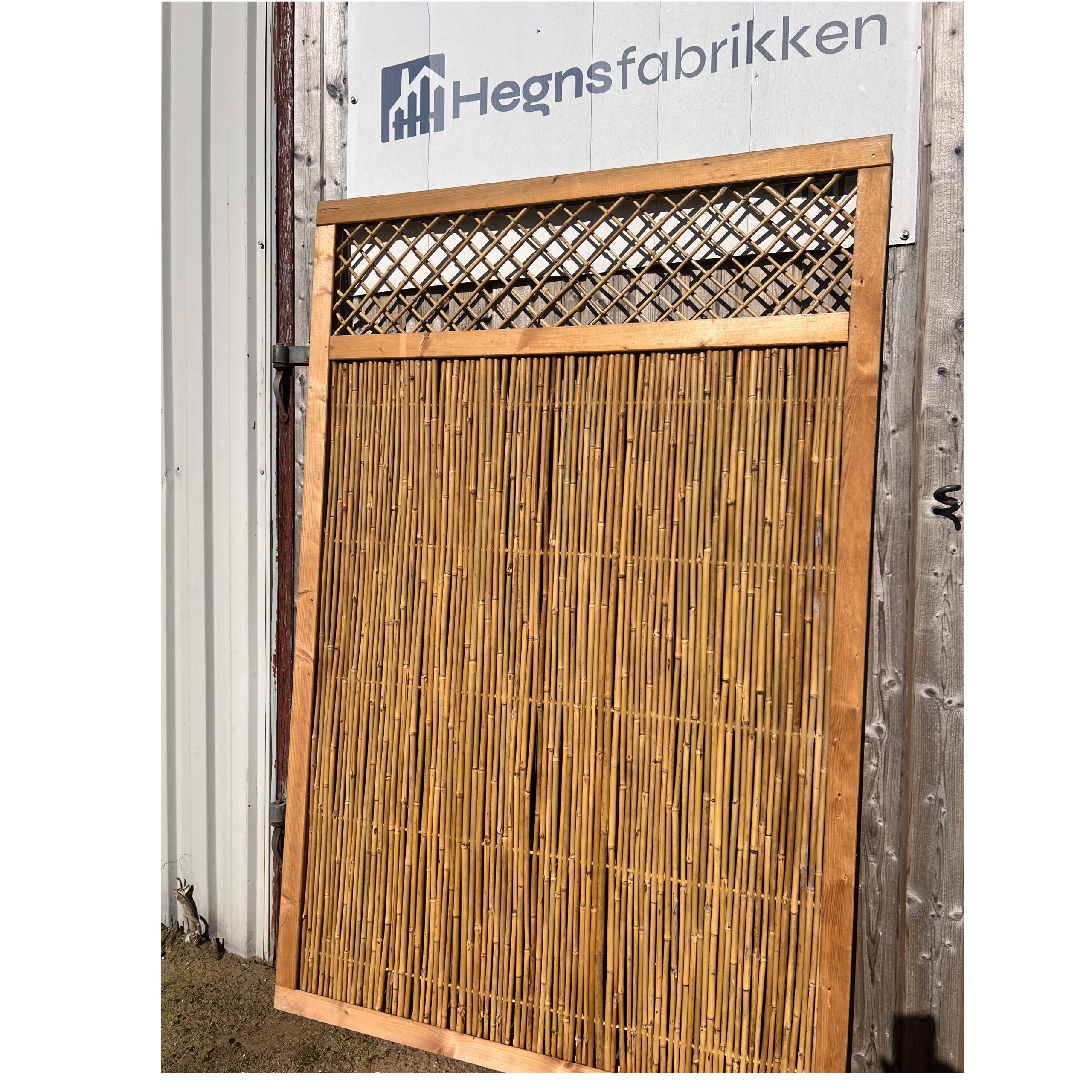 En Bambus hegn 110x170 cm træramme med en elegant gittertop læner sig op ad en trævæg under "Hegnsfabrikken"-skiltet i et lyst, solbeskinnet udendørs miljø.