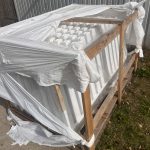 En trækasse pakket ind i iturevet hvid plastik står på beton og græs. Indeni er der hvide paneler - sandsynligvis Outlet: Stakithegn i plast - Profexline 180x80 cm bue op. "RETUR" og "RETURN" er markeret på kassens træramme.