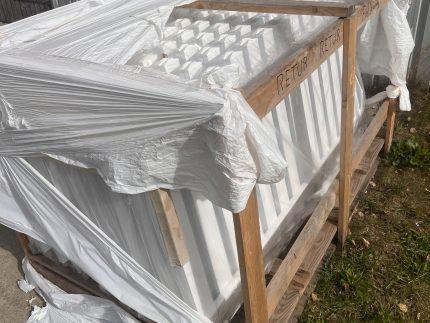 En trækasse pakket ind i iturevet hvid plastik står på beton og græs. Indeni er der hvide paneler - sandsynligvis Outlet: Stakithegn i plast - Profexline 180x80 cm bue op. "RETUR" og "RETURN" er markeret på kassens træramme.
