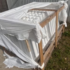 En trækasse pakket ind i iturevet hvid plastik står på beton og græs. Indeni er der hvide paneler - sandsynligvis Outlet: Stakithegn i plast - Profexline 180x80 cm bue op. "RETUR" og "RETURN" er markeret på kassens træramme.