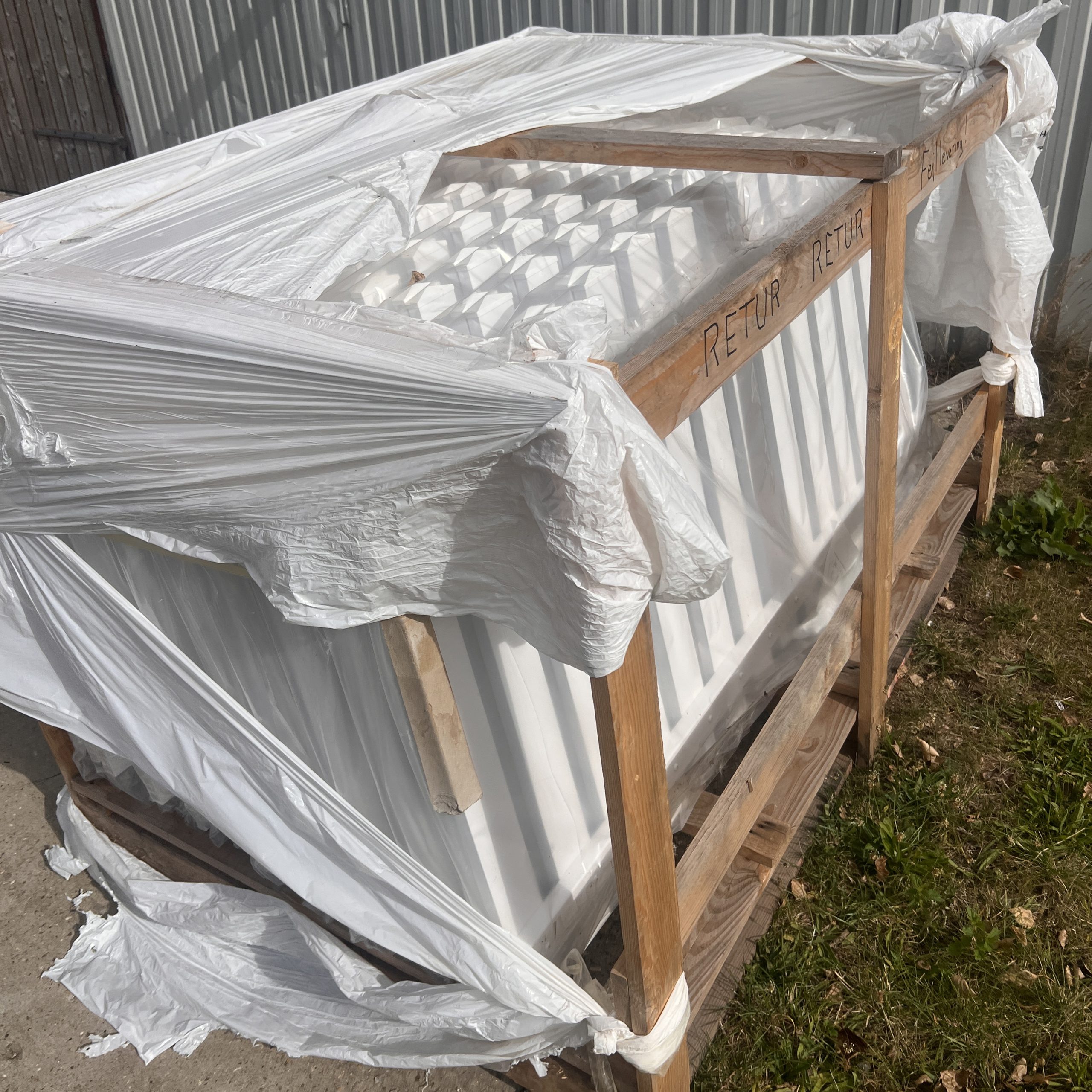 En trækasse pakket ind i iturevet hvid plastik står på beton og græs. Indeni er der hvide paneler - sandsynligvis Outlet: Stakithegn i plast - Profexline 180x80 cm bue op. "RETUR" og "RETURN" er markeret på kassens træramme.