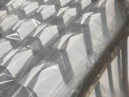 Stakke af hvid Outlet: Stakithegn i plast - Profexline 180x80 cm bue op er pakket ind i klar plast, og deres pyramideformede toppe kaster kantede skygger og mønstre i sollyset.