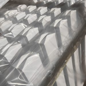 Stakke af hvid Outlet: Stakithegn i plast - Profexline 180x80 cm bue op er pakket ind i klar plast, og deres pyramideformede toppe kaster kantede skygger og mønstre i sollyset.