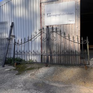 En dekorativ sort smedejernsport læner sig op ad en bygning i bølgeblik med et stort skilt, hvor der står "Hegnsfabrikken Hal 2". Sollyset kaster skygger på jorden og bygningens væg.