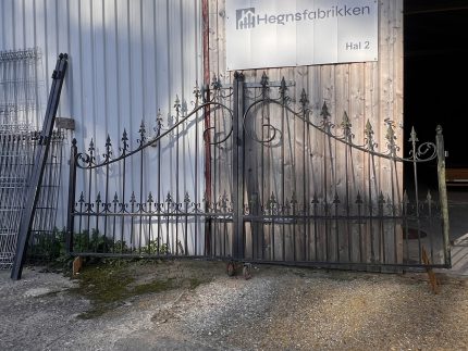 En dekorativ sort smedejernsport læner sig op ad en bygning i bølgeblik med et stort skilt, hvor der står "Hegnsfabrikken Hal 2". Sollyset kaster skygger på jorden og bygningens væg.