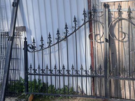 Sort smedejernslåge med dekorative fleur-de-lis-spidser læner sig op ad en bølgepapvæg. Porten er delvist åben med hjul i bunden, og der vokser græs i nærheden.