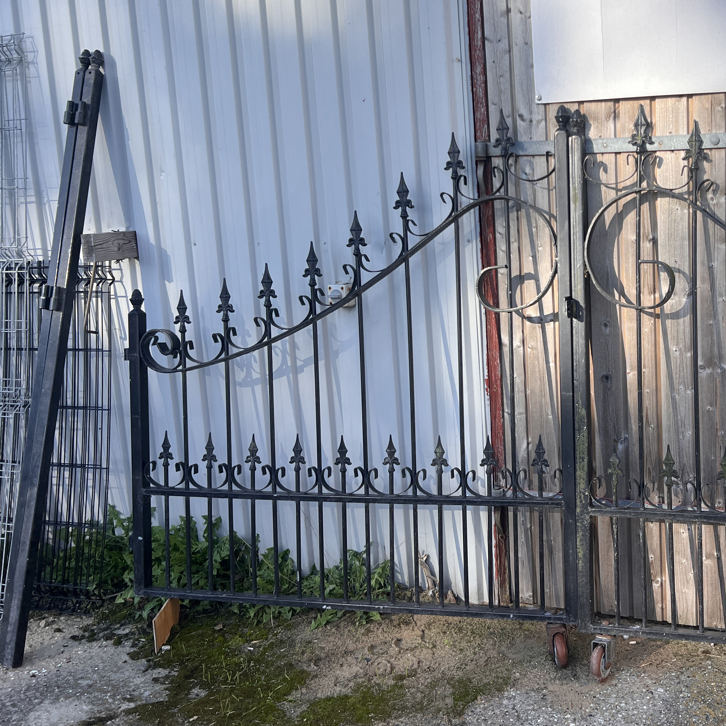 Sort smedejernslåge med dekorative fleur-de-lis-spidser læner sig op ad en bølgepapvæg. Porten er delvist åben med hjul i bunden, og der vokser græs i nærheden.