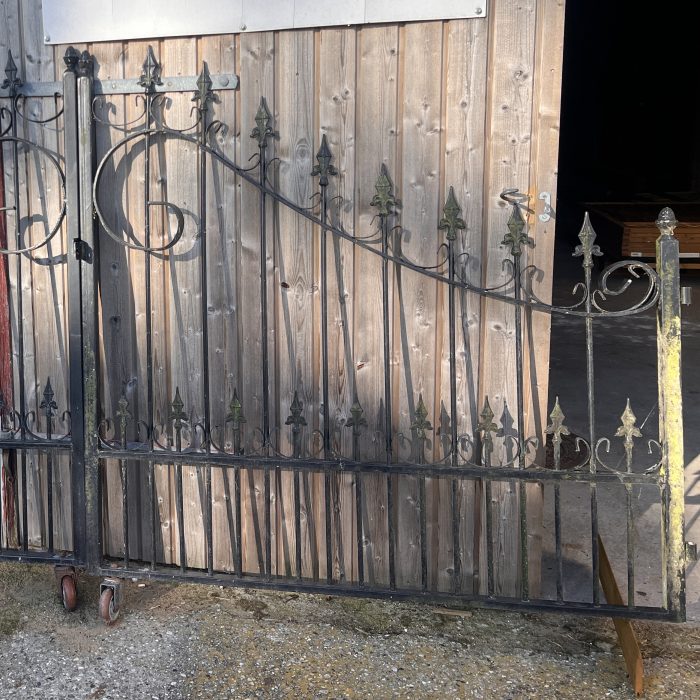 En dekorativ smedejernslåge med spidse finials læner sig op ad en trævæg. Porten har udsmykket rulleværk og står på små røde hjul forneden. Baggrunden er en trækonstruktion med en delvist åben dør.