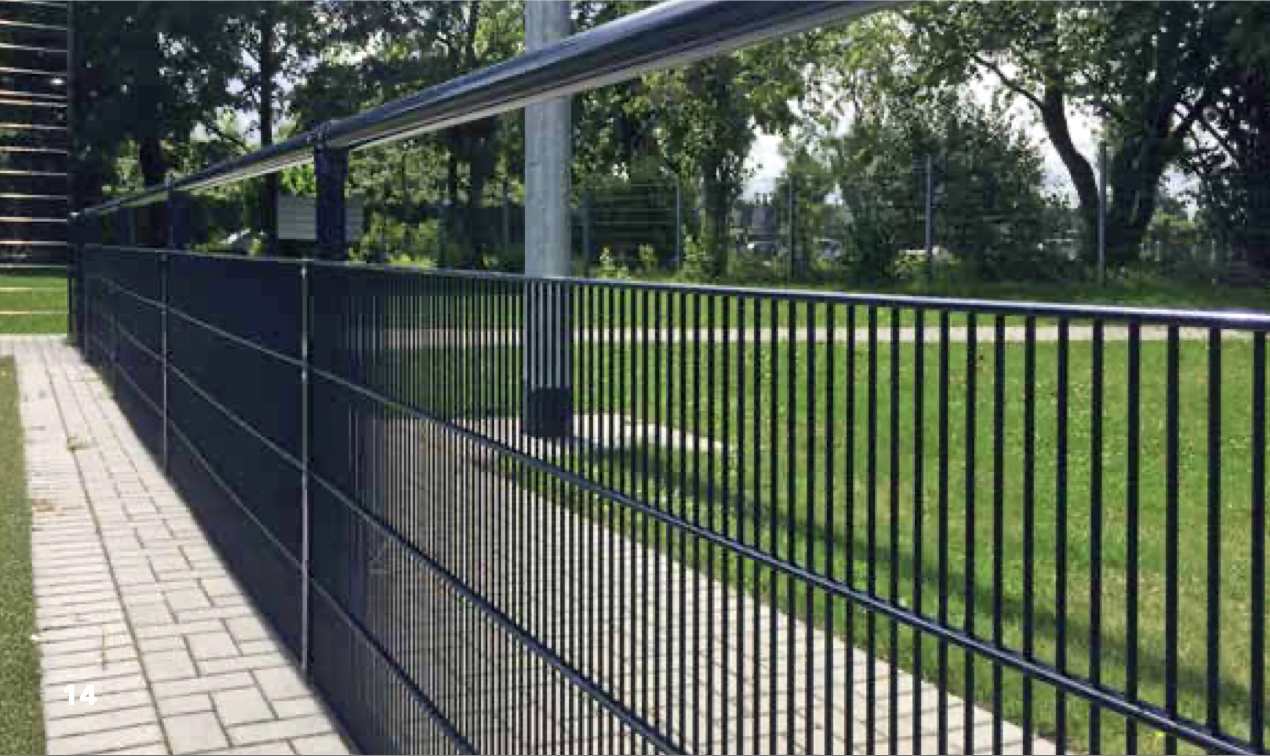 A Rørhegn med Panelfyld - Varmgalvaniseret eller Pulverlakeret (flere RAL-farver) løber parallelt med en brolagt gangsti og adskiller et græsareal fra stien med træer og grønt i baggrunden under en delvist overskyet himmel.