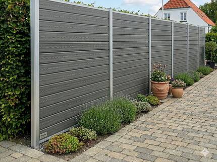En Komposithegn med aluminiumsstolper & alu top/bundskinner - Type 3 i moderne grå står ved siden af en brolagt gangsti med potteplanter og grønne planter, sat op mod et hvidt hus med rødt tag og flag under en blå himmel.
