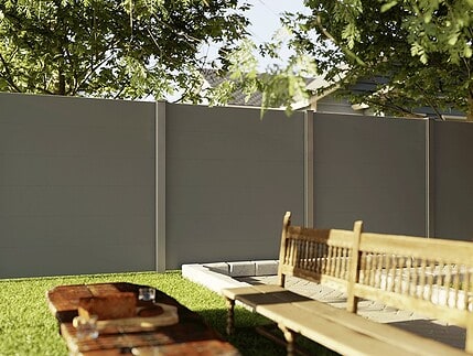 En træbænk og et picnicbord står på en græsplæne ved siden af et moderne, gråt hegn, der skærmer for indkig, mens løvfældende træer giver skygge, og sollyset siver ind gennem grenene.