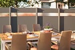 Et udendørs spisebord med beige stole, dækket med tallerkener og glas, står på en terrasse foran et gråt hegn med frostede paneler, der skærmer for indkig. På bordet står en potteplante med lilla blomster, og i baggrunden ses et hus og træer.