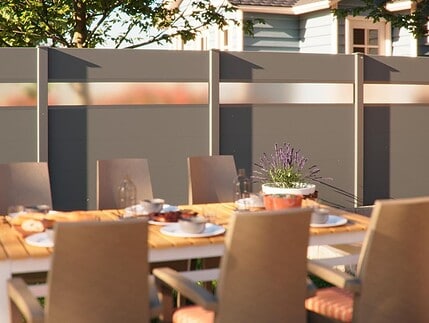 Et udendørs spisebord med beige stole, dækket med tallerkener og glas, står på en terrasse foran et gråt hegn med frostede paneler, der skærmer for indkig. På bordet står en potteplante med lilla blomster, og i baggrunden ses et hus og træer.