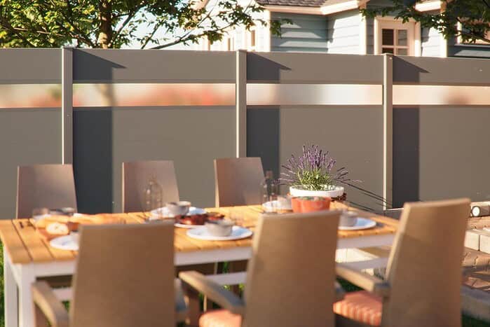 Et udendørs spisebord med beige stole, dækket med tallerkener og glas, står på en terrasse foran et gråt hegn med frostede paneler, der skærmer for indkig. På bordet står en potteplante med lilla blomster, og i baggrunden ses et hus og træer.