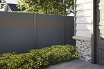 Et gråt hegn, der skærmer for indkig, med en vandret brun stribe står ved siden af et hus med hvidt yderbeklædning og detaljer i sten. Gule blomstrende planter vokser langs hegnets fod under løvrige træer.