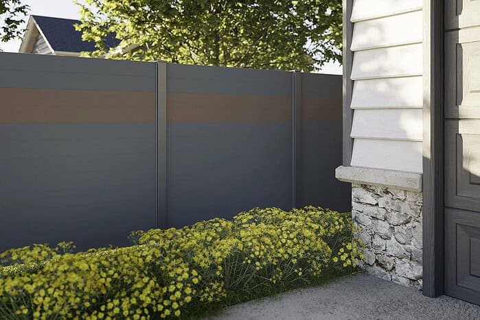 Et gråt hegn, der skærmer for indkig, med en vandret brun stribe står ved siden af et hus med hvidt yderbeklædning og detaljer i sten. Gule blomstrende planter vokser langs hegnets fod under løvrige træer.