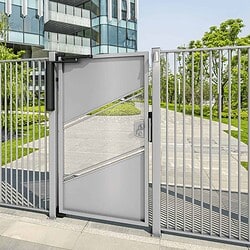 En moderne fodgængerport i metal med et geometrisk mønster står lukket, omgivet af lodrette stænger. Bag den fører en sti forbi grønne græsplæner og træer mod en moderne glasbygning under en klar himmel.