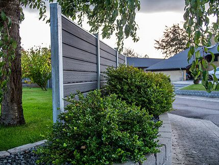 Et moderne gråt hegn med grønne buske og et træ i forgrunden, ved siden af en asfalteret indkørsel og bolighuse i baggrunden.