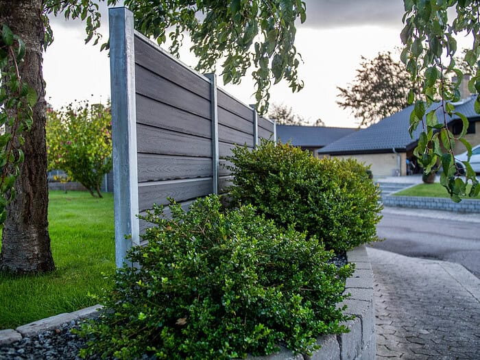 Et moderne gråt hegn med grønne buske og et træ i forgrunden, ved siden af en asfalteret indkørsel og bolighuse i baggrunden.