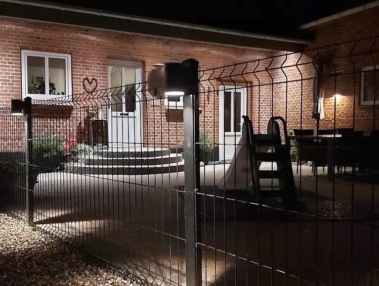 3D-panelhegn med lys i stolperne foran et murstenshus om aftenen