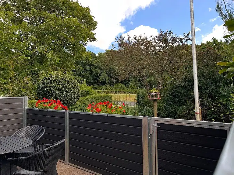 Komposithegn om en terrasse med havemøbler og røde pelargonier