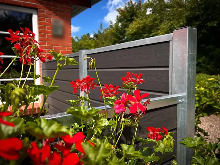 Røde pelargonier foran komposithegn med varmgalvaniserede stolper